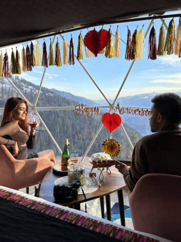 A couple celebrating love with a glass of wine, cake and breathtaking views from their luxury dome at GlampEco Manali